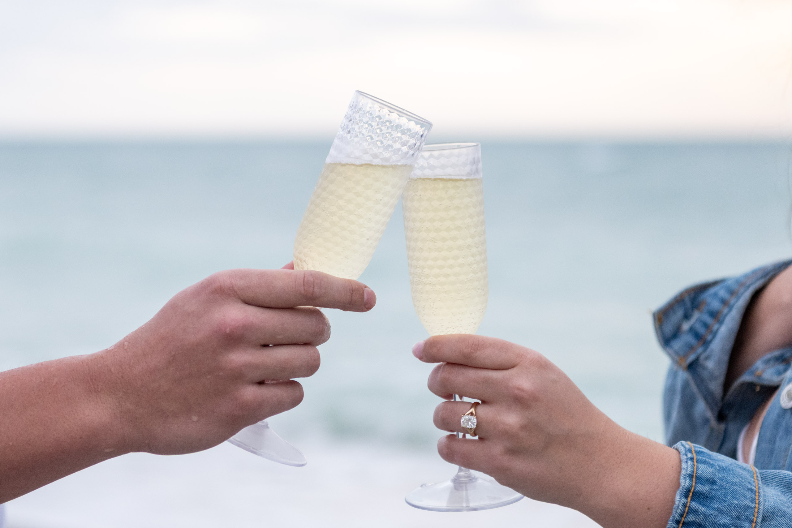 couple toast with champagne on the beach and show off her engagement ring