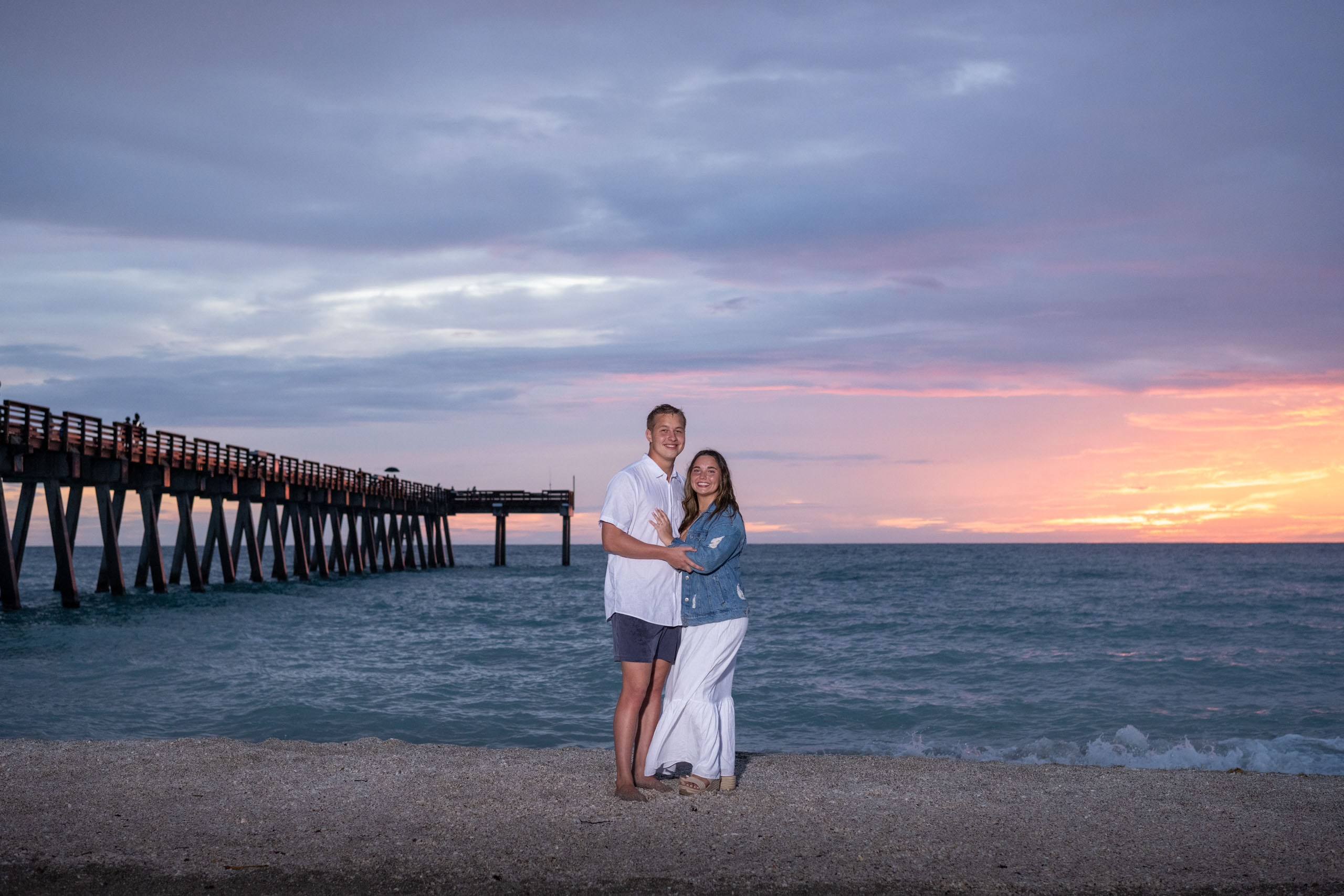 engaged couple smile by the Venice Fishing Pier as the sky turns pink after a storm