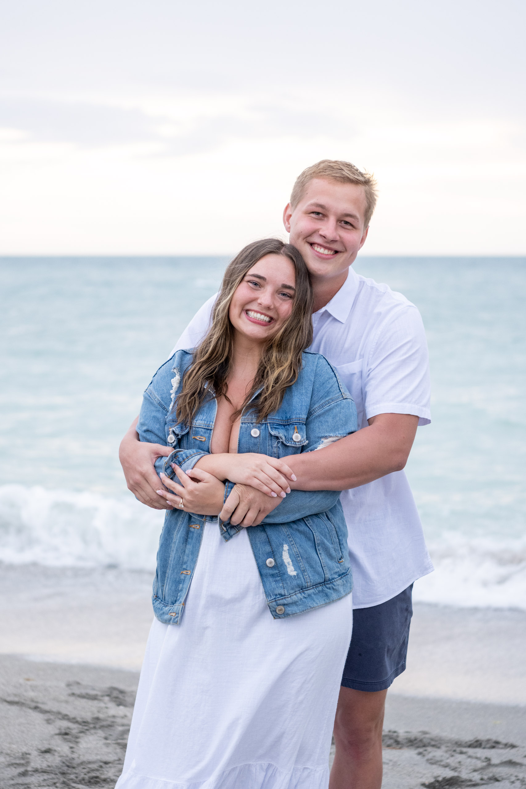 young engaged couple smile together as he holds her on the beach