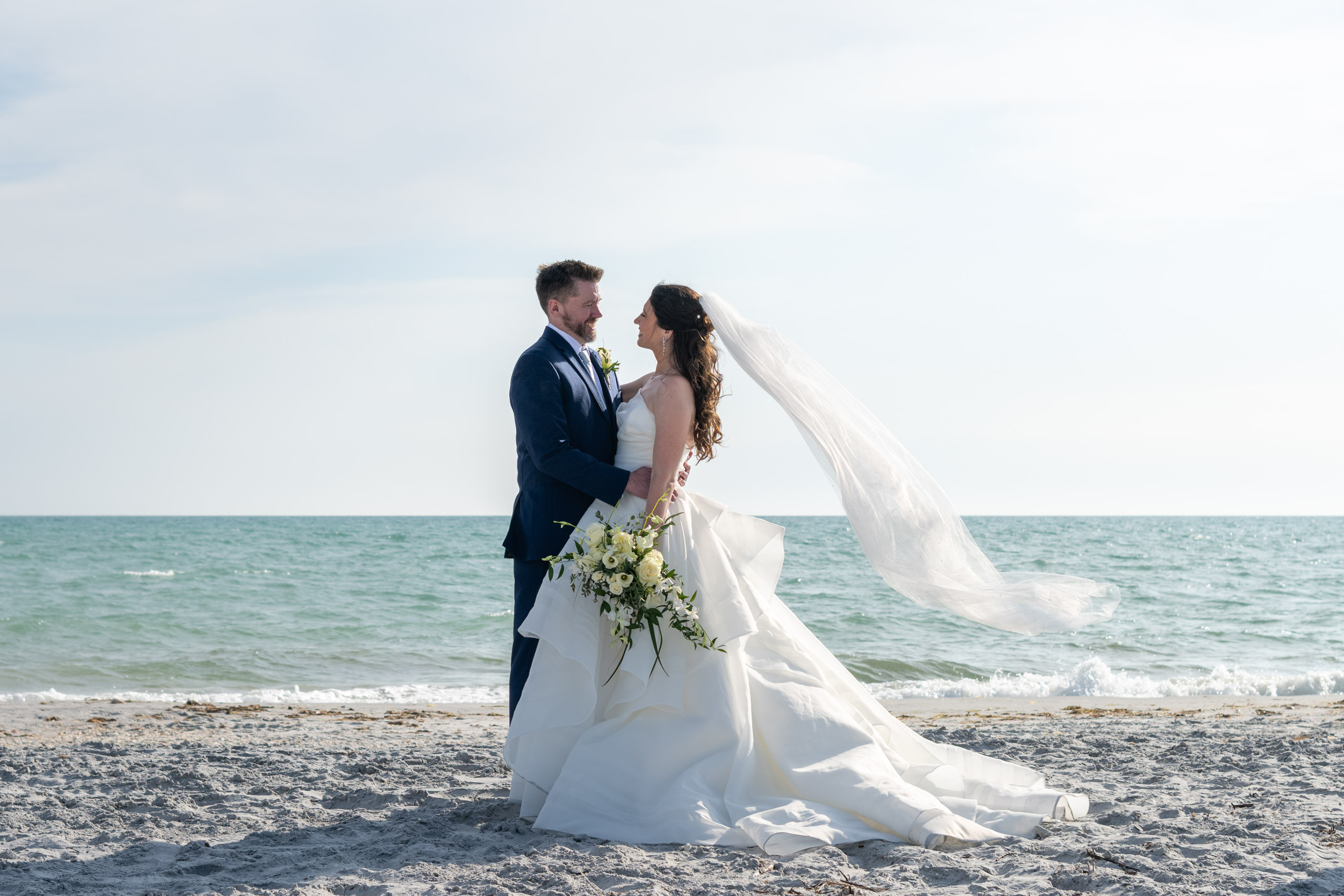 bride and groom looking at each other on the beach with her veil blowing behind her