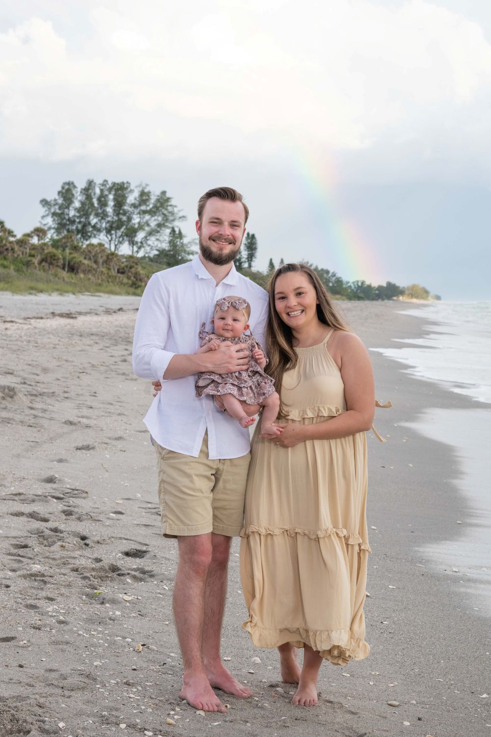 parents hold their baby girl on the beach with a rainbow in the background