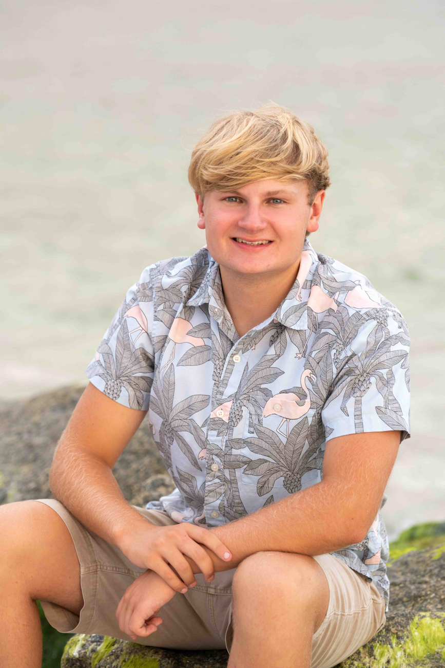 senior boy smiling as he sits on a rock with the water behind him