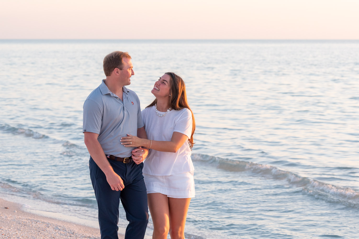 engagement session at sunset as couple walk along beach looking lovingly at each other