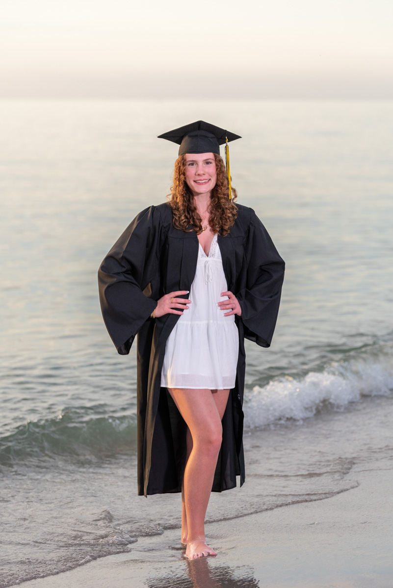 senior girl in black cap and gown standing on the beach