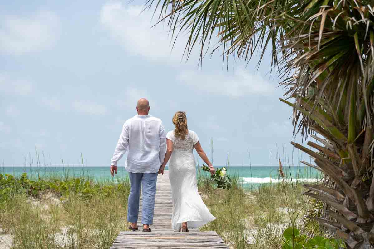 bride and groom walk towards the beach and Gulf while holding hands