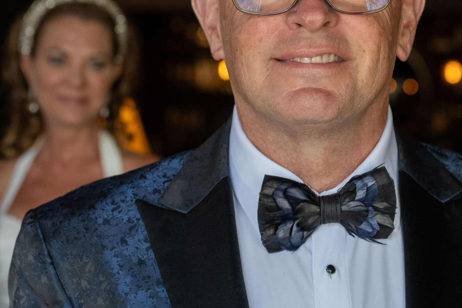 close up of a dark blue feather bowtie worn by the groom with his bride looking on from the background