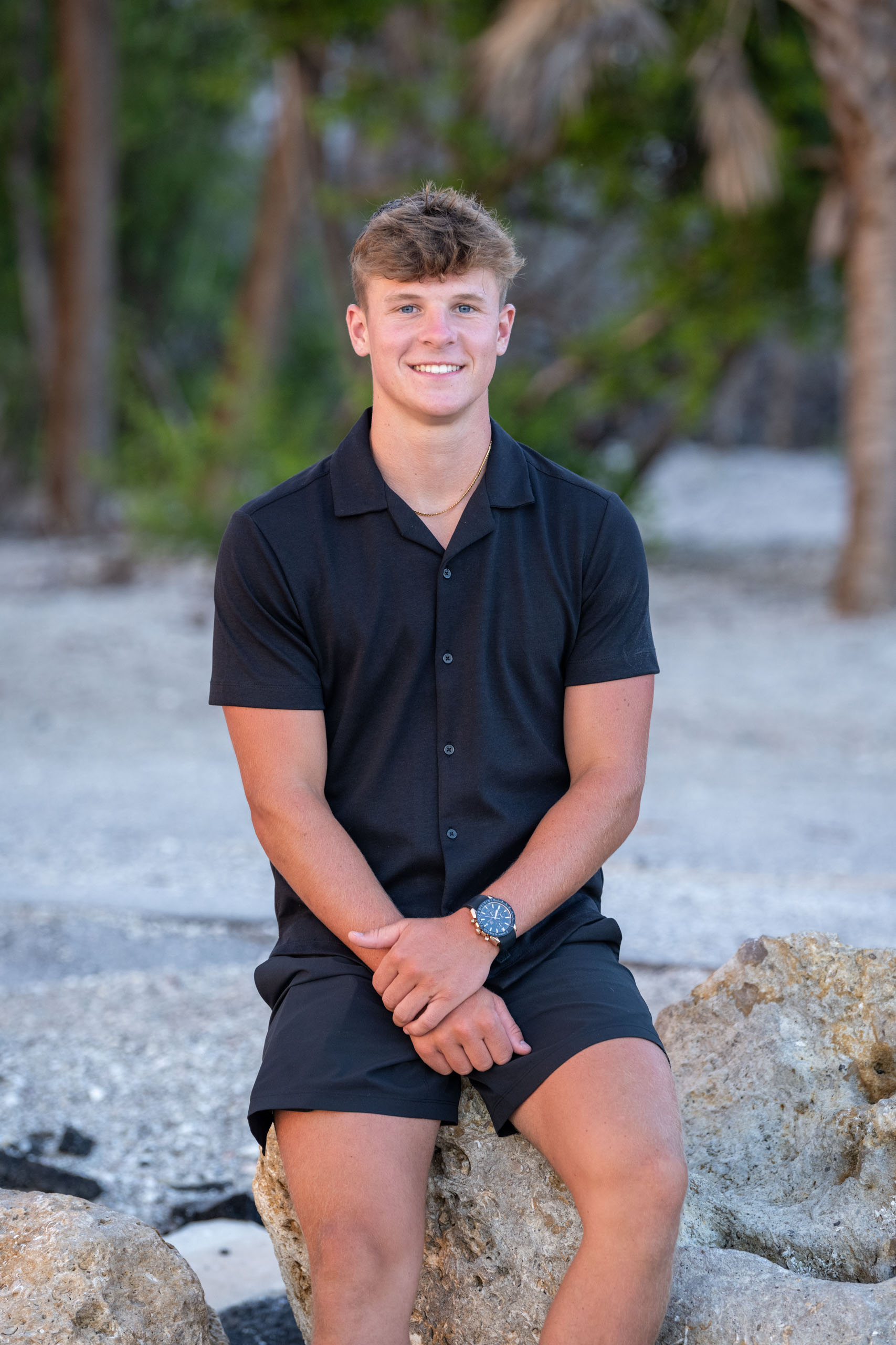 senior boy dressed in black top and shorts sitting on rocks