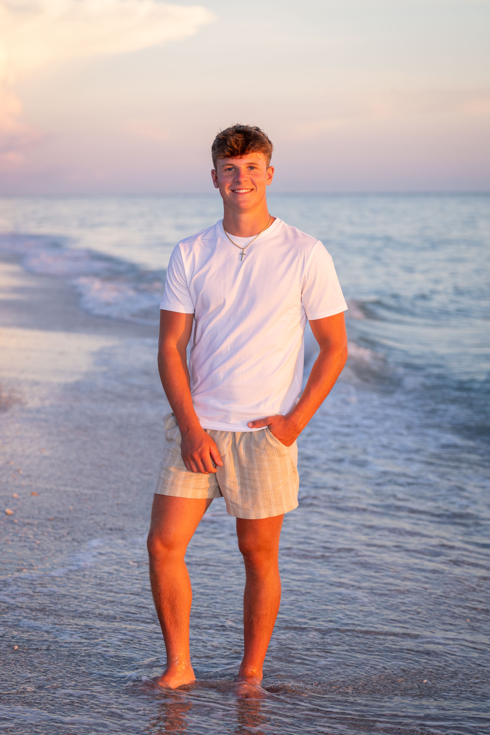 senior boy on beach at sunset. He's in a white t shirt and tan shorts