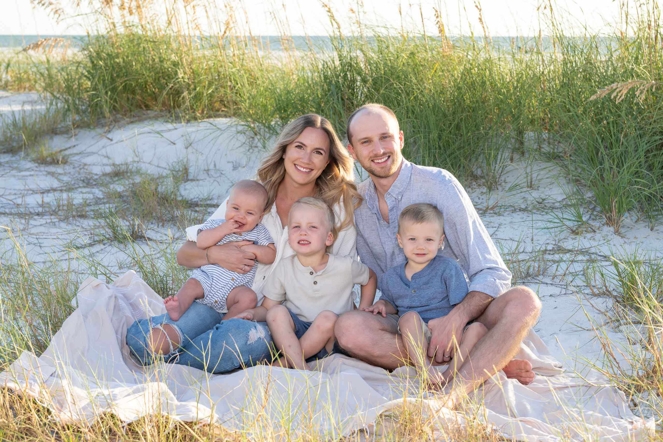 family sits on a beach blanket with their three young children smiling