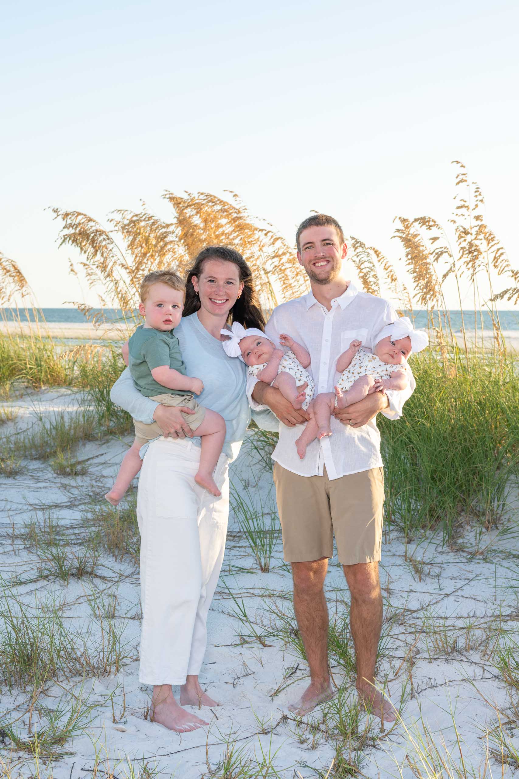 parents hold their son and newborn twins on the beach with seagrass and the Gulf behind them