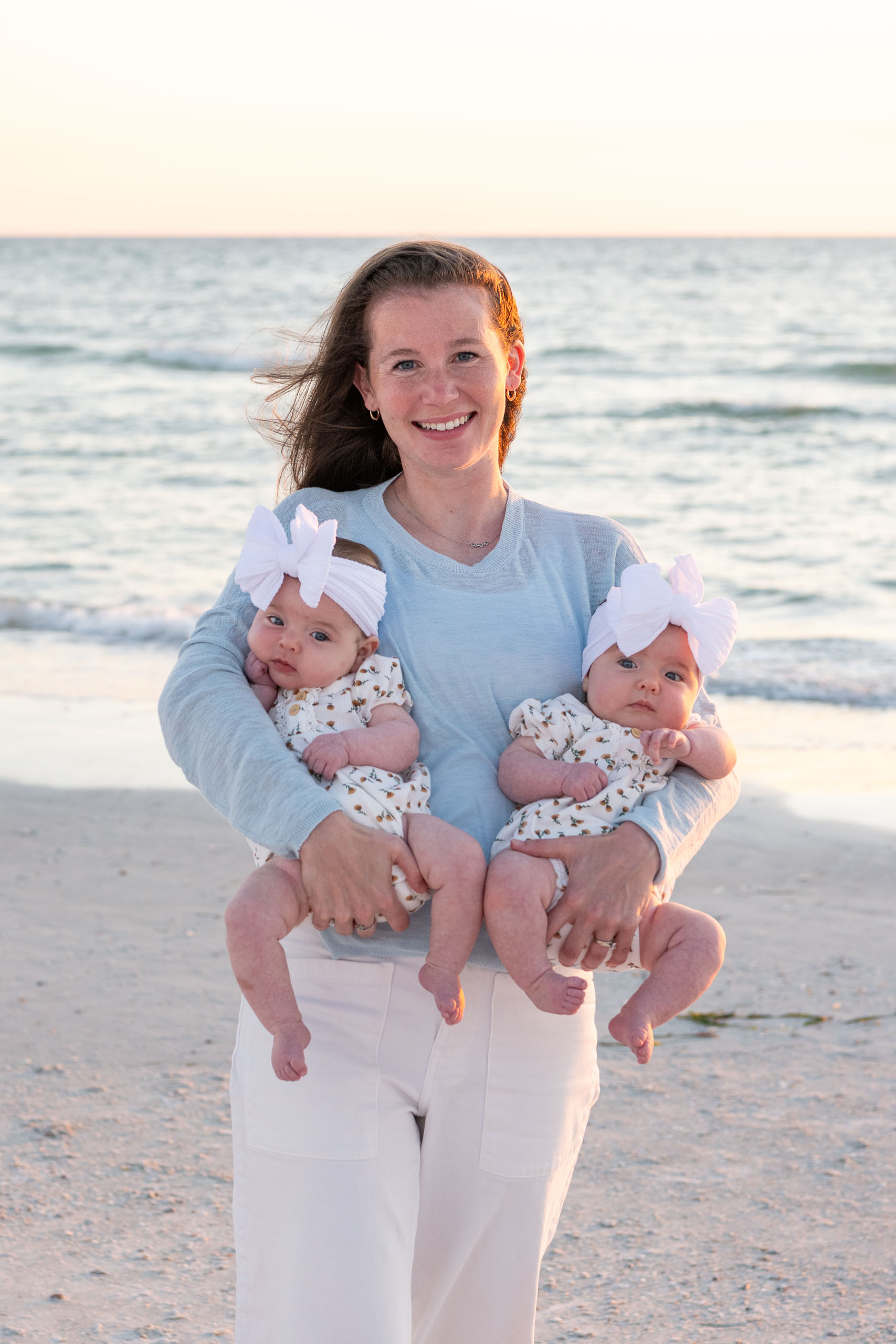 mom holds her twin girls on the beach with the calm water behind her