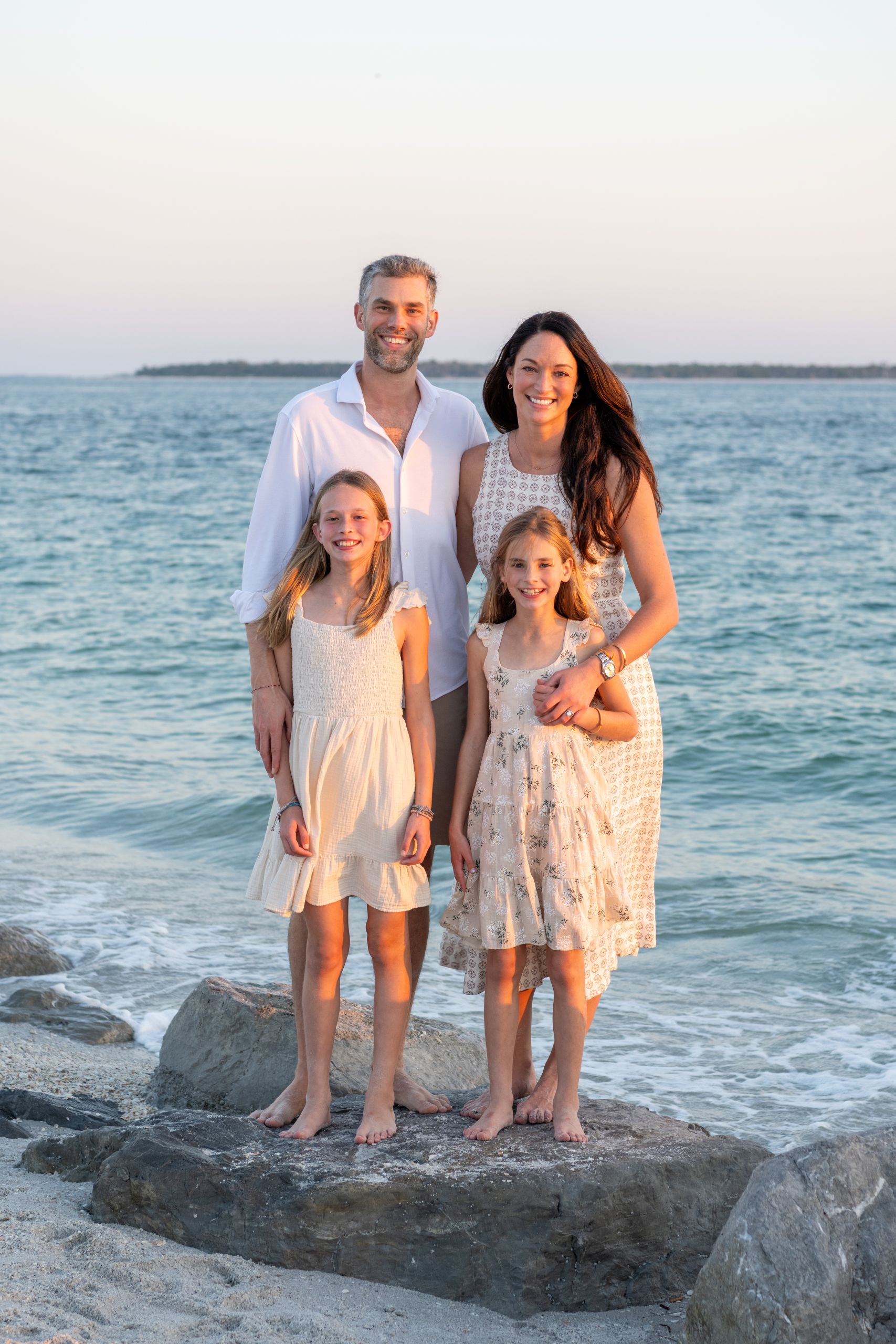 mom and dad wrap their arms around their daughters as they stand on a rock with the water behind them