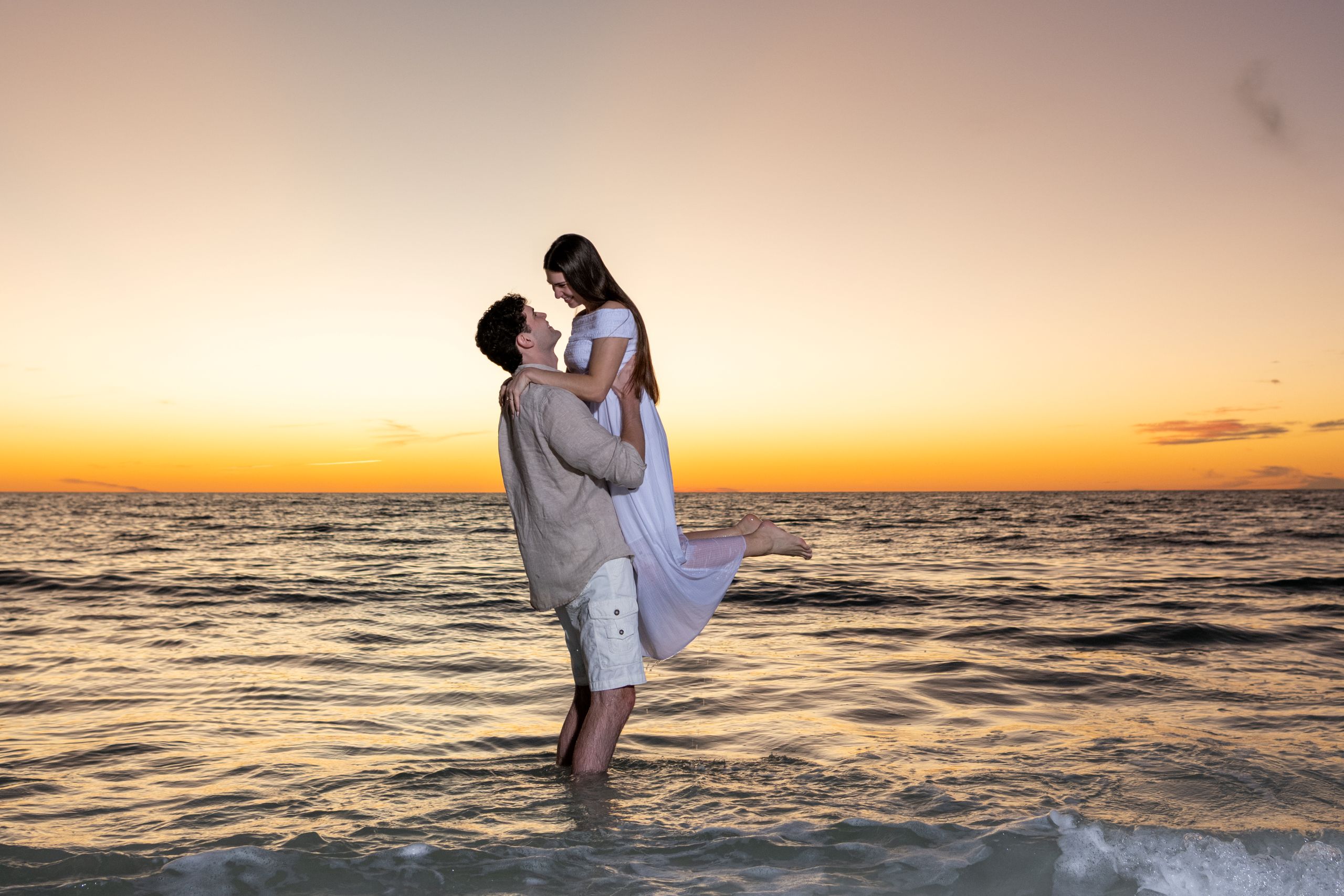 couple standing in the water and the guy lifts his girlfriend up after sunset