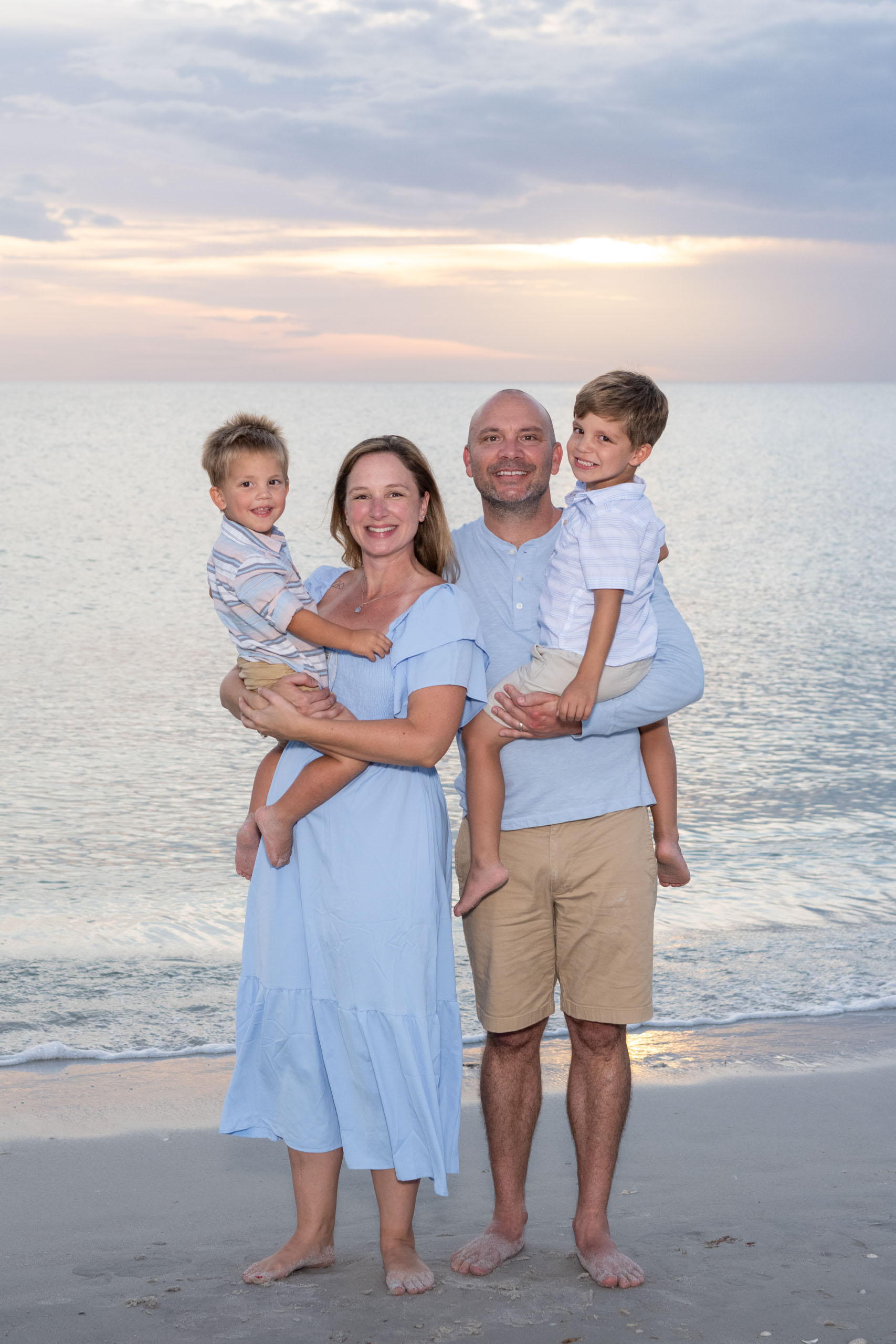 parents hold their smiling sons on the beach with the golden hour glow of sunset behind them