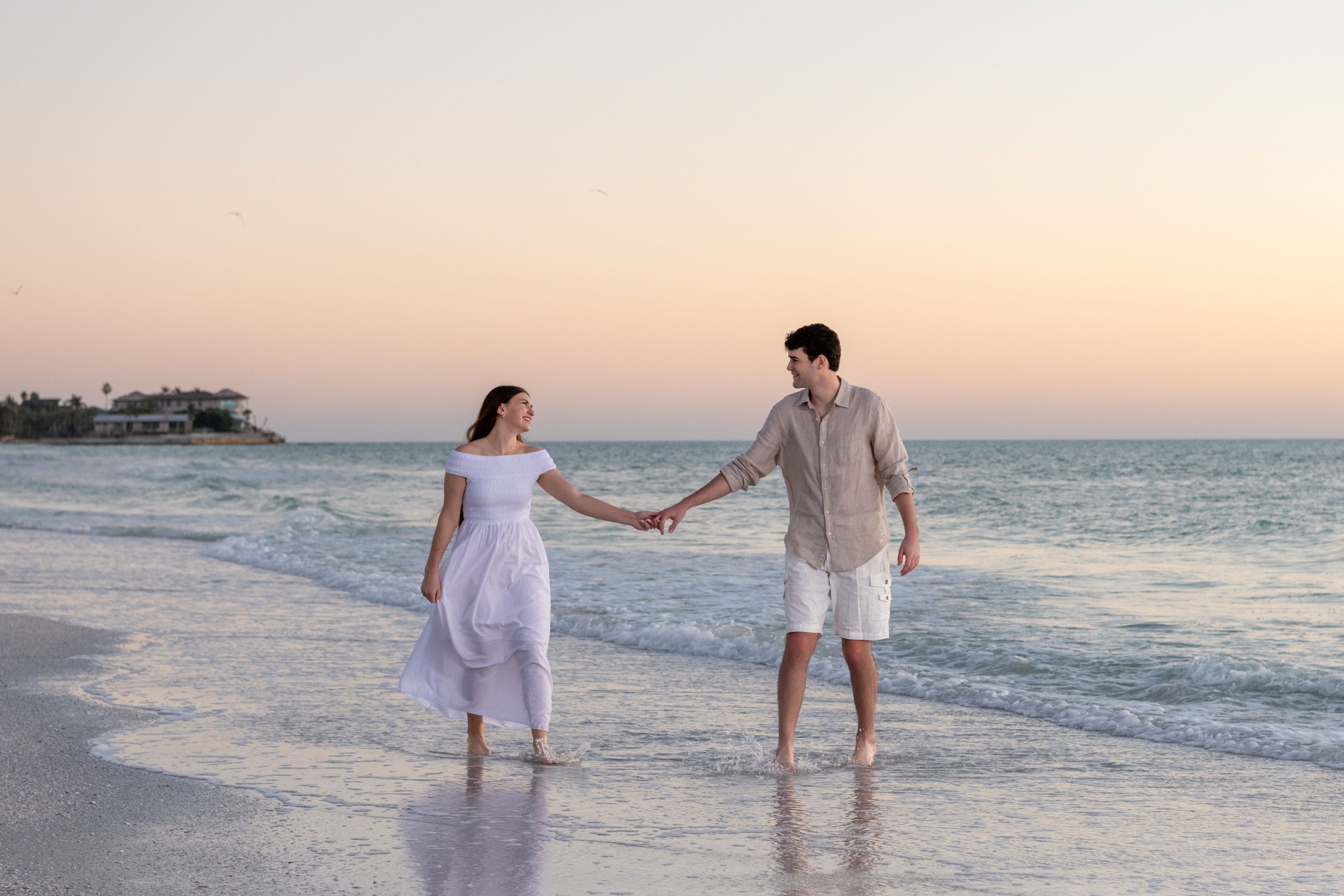 girl holds her fiance's hand as she looks lovingly in his eyes as they walk along the beach at sunset