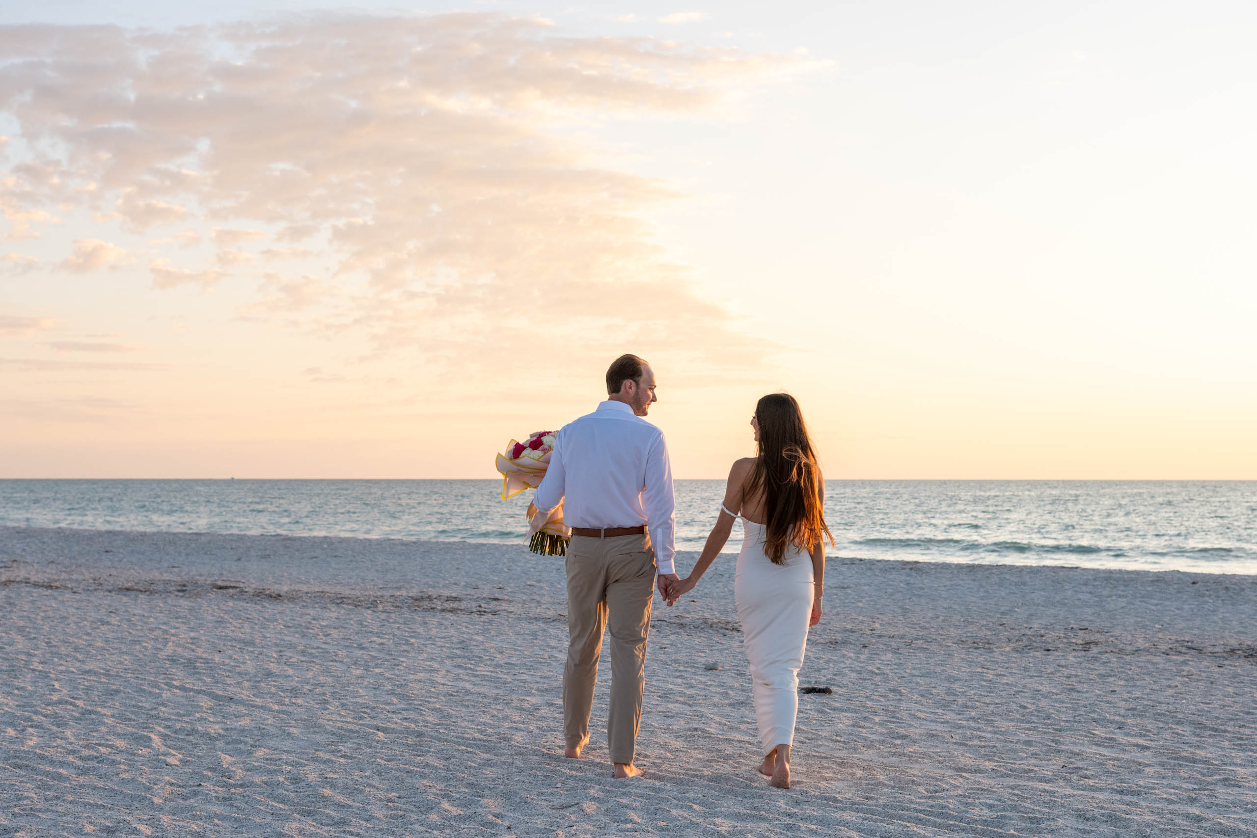 couple walk casually towards the water with the guy holding the bouquet of roses