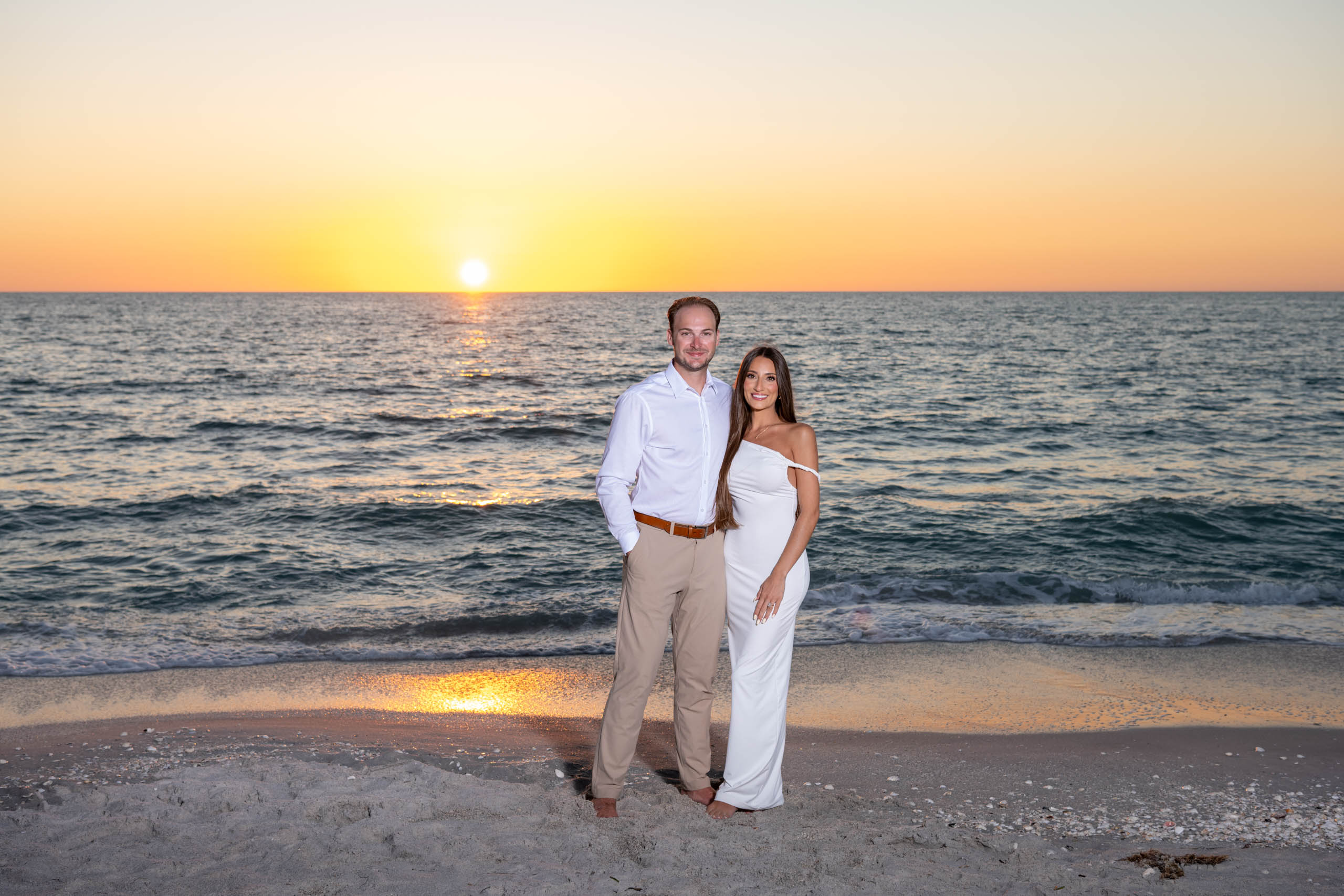 couple stand together on the beach as the sun sets behind them