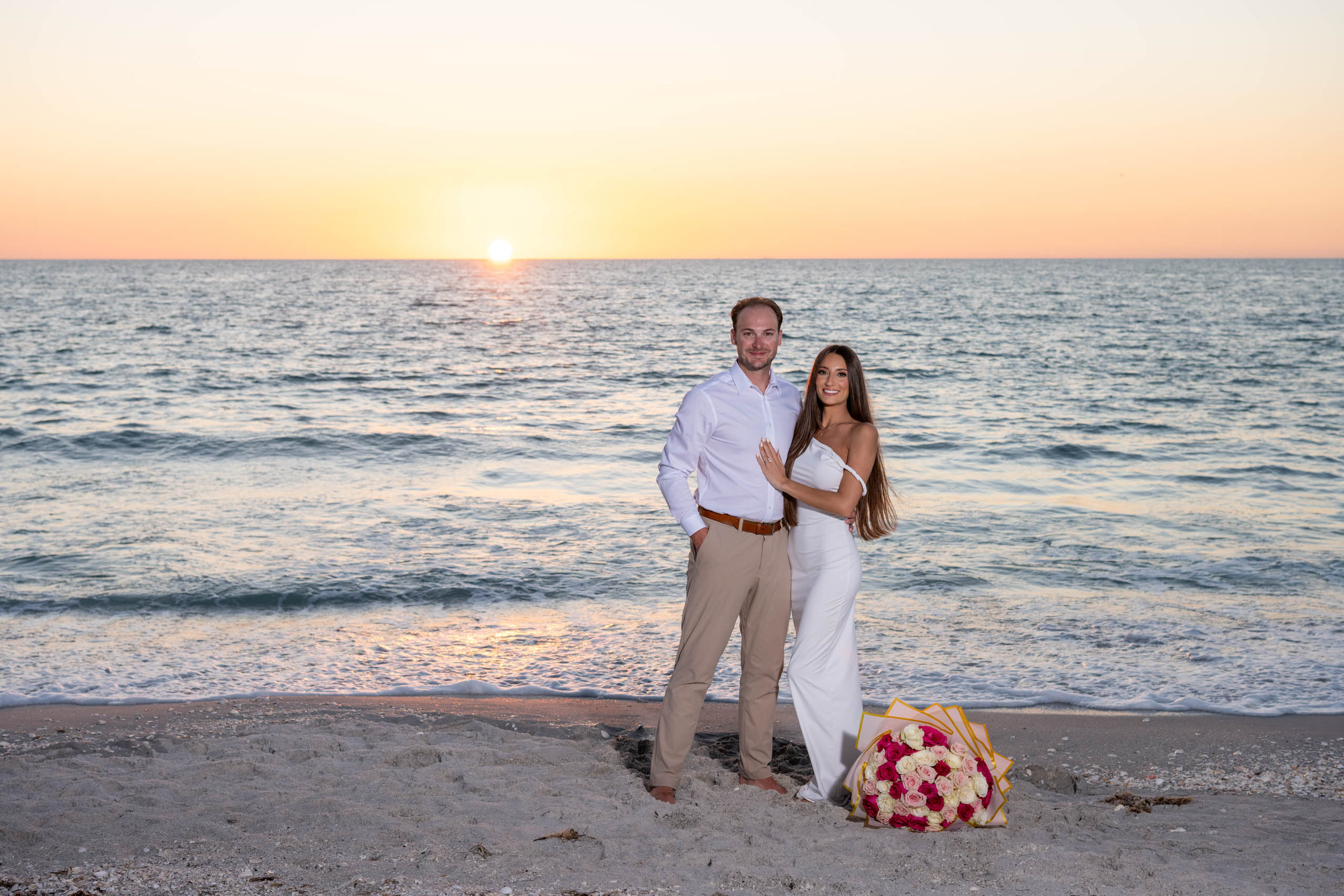 engaged couple show off her large bouquet of roses on the beach at sunset