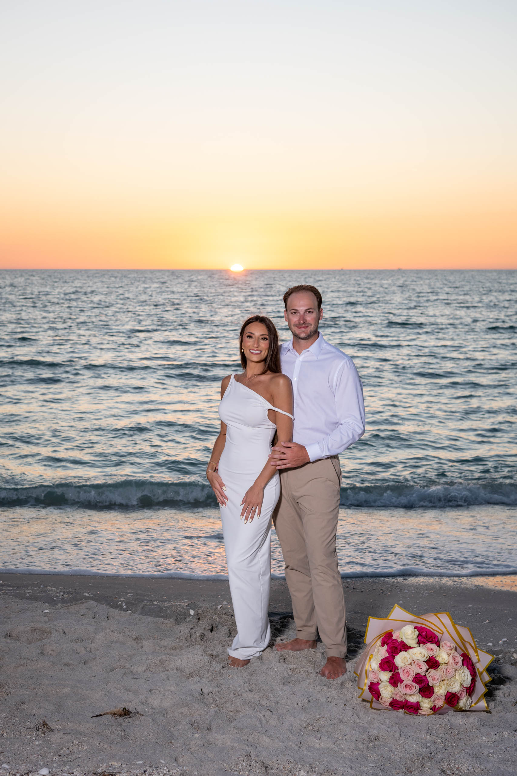 newly engaged couple dressed in white pose with a bouquet of roses in the sand as the sun sets behind them