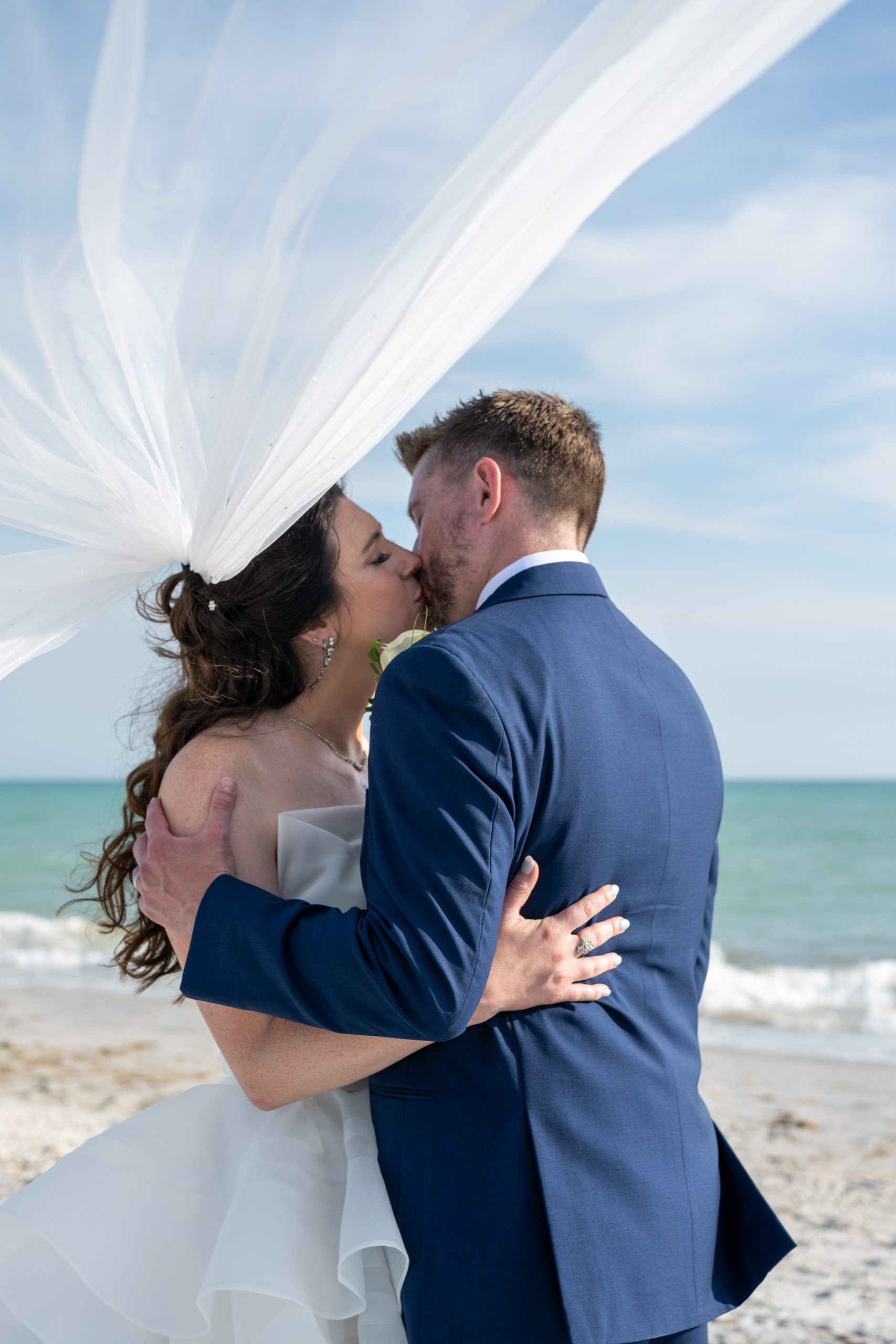 bride and groom kiss under her veil as they stand on the beach
